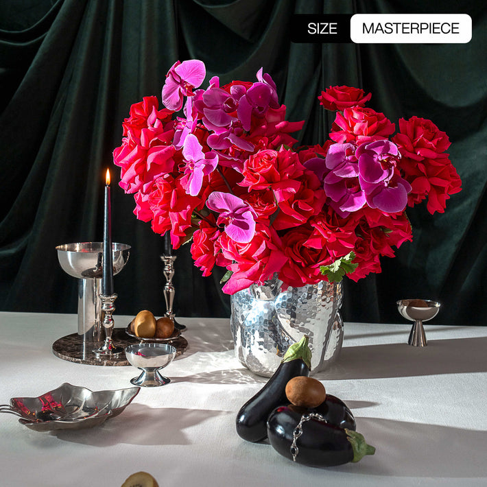 Decorative table setting with pink Floyd roses and pink phalaenopsis orchids, fruits, and silverware against a dark curtain.