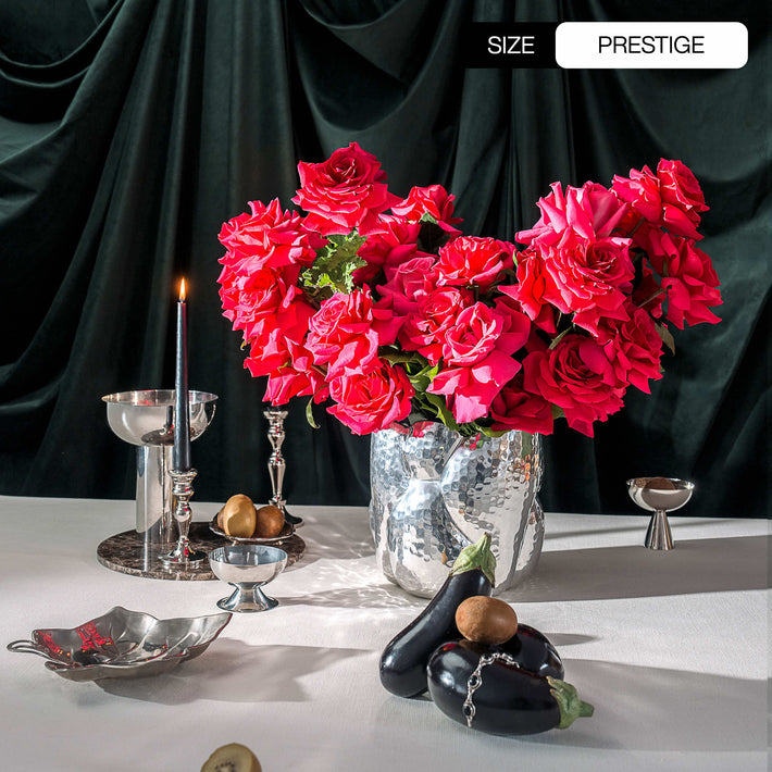 Decorative table setting with pink Floyd roses and pink phalaenopsis orchids, fruits, and silverware against a dark curtain.
